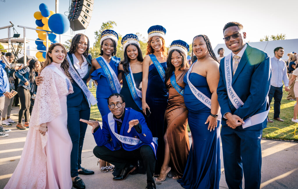 A group of diverse students standing outside facing the camera and wearing formal attire with blue and white Homecoming Royal Court sashes. 