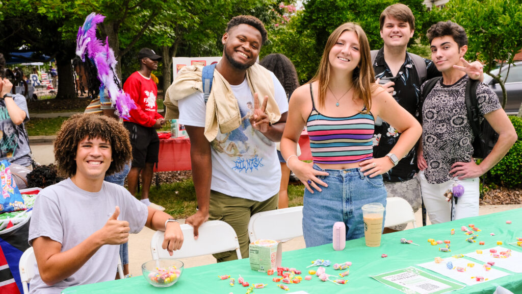 A diverse group of students smiling at the camera and standing at a table outside.